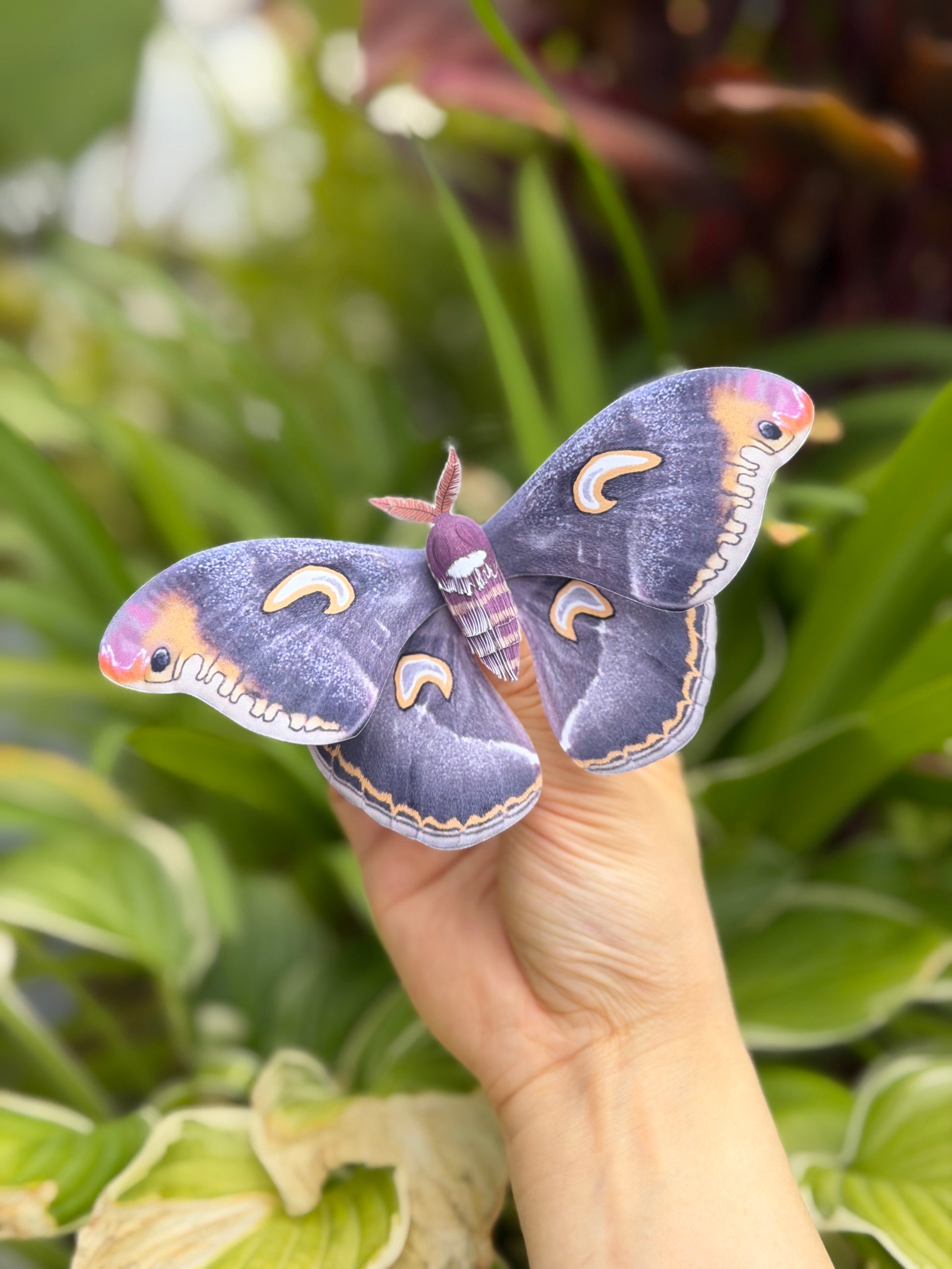 Hand holding a Dark Moon Emperor Moth made of PAPER with a blurred green plant background