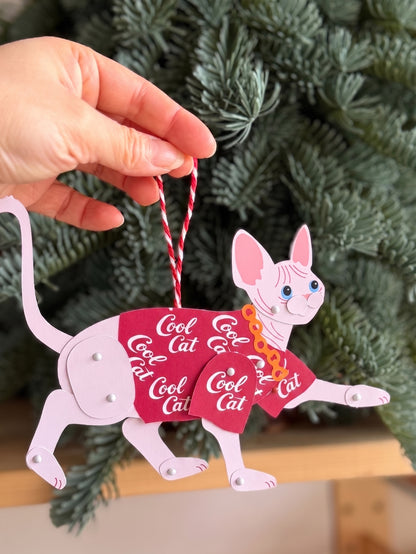Decorative ornament Sphynx in Red Shirt Paper Doll, held against a Christmas tree.